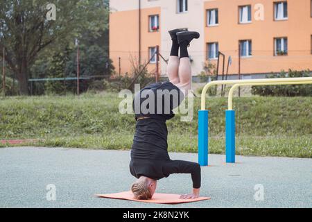 Der Calisthenics-Athlet führt auf einem Outdoor-Workout-Platz auf einer orangefarbenen Matte einen Kopfstand mit solidem Kern und armer Stabilität aus. Die mittlere Phase des e Stockfoto