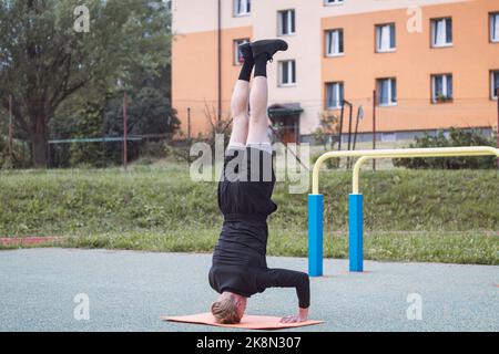 Der Calisthenics-Athlet führt auf einem Outdoor-Workout-Platz auf einer orangefarbenen Matte einen Kopfstand mit solidem Kern und armer Stabilität aus. Die letzte Phase des Ex Stockfoto