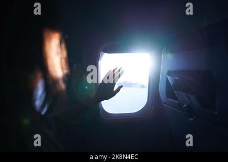 Weibliche Hand berührt die warmen Sonnenstrahlen vom Flugzeugfenster. Frau freut sich auf die Reise in ihren Urlaub. Mädchen auf einer Reise Flug sitzen in Stockfoto