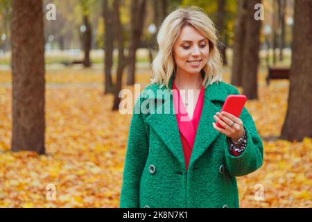 Blonde Frau mittleren Alters lächelt und benutzt Smartphone im herbstlichen Stadtpark. Stilvoll gekleidete Frau in rosa Anzug und warmen grünen Mantel. Rosa Smartphone Stockfoto