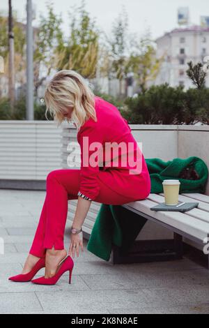 Geschäftsfrau in pinkem Anzug, die aufgrund der hohen Absätzen an Schuhen Schmerzen in den Beinen leidet und unbequeme rosa Schuhe auszieht. Stockfoto