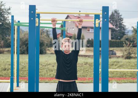 Der zielorientierte blonde Junge baut im Sommer die Kraft seines eigenen Körpers auf dem Trainingsfeld auf. Training von Trizeps, Bizeps, Schultern und Bauch. Stockfoto