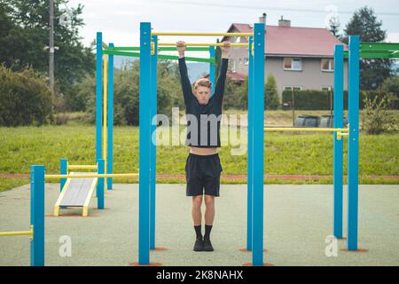 Der zielorientierte blonde Junge baut im Sommer die Kraft seines eigenen Körpers auf dem Trainingsfeld auf. Training von Trizeps, Bizeps, Schultern und Bauch. Stockfoto