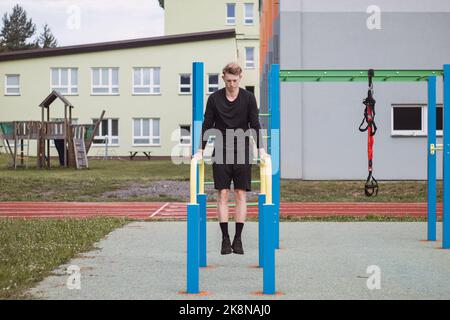 Der zielorientierte blonde Junge baut im Sommer die Kraft seines eigenen Körpers auf dem Trainingsfeld auf. Training von Trizeps, Bizeps, Schultern und Bauch. Stockfoto