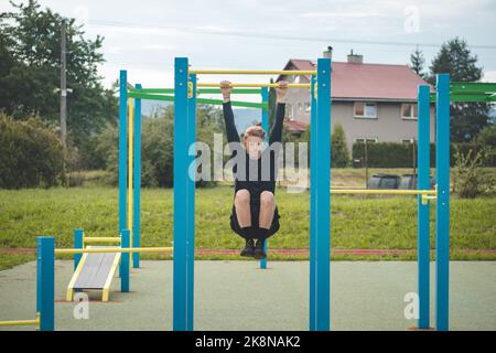 Der zielorientierte blonde Junge baut im Sommer die Kraft seines eigenen Körpers auf dem Trainingsfeld auf. Training von Trizeps, Bizeps, Schultern und Bauch. Stockfoto