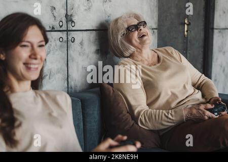 Porträt einer lachenden älteren Frau mit ihrer erwachsenen Tochter, die zu Hause Videospiele spielt und auf dem Sofa sitzt. Technologie und Lifestyle Stockfoto