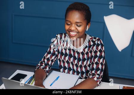 Porträt einer lächelnden jungen afroamerikanischen Frau, die an einem Laptop arbeitet, während sie an einem Tisch im modernen Bürogebäude sitzt. Sitzen am Schreibtisch mit Gadgets Stockfoto