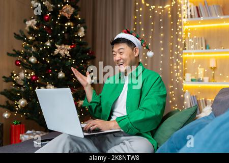 Fröhlicher Mann in weihnachtshut und grünem Hemd, der mit Freunden über Videoanruf spricht, asiatischer Mann, der Weihnachten und Neujahr zu Hause feiert, sitzt auf dem Sofa im Wohnzimmer mit Laptop. Stockfoto