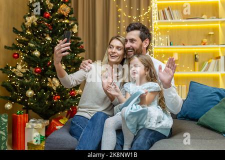 Glückliche Familie feiert Neujahr und weihnachten, Ehemann, Ehefrau und kleine Tochter sitzen auf dem Sofa im Wohnzimmer und sprechen mit Verwandten in Videoanruf, Telefon für die Fernkommunikation. Stockfoto