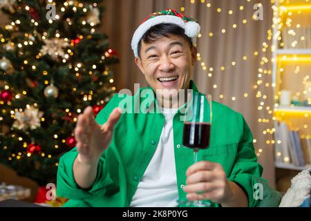 Fröhlicher asiatischer Mann an weihnachten und Neujahr begrüßt Freunde, Mann spricht bei einem Videoanruf mit Kollegen, die zu Hause auf dem Sofa sitzen, mit Hut und einem Glas Wein, Blick auf das Internet, Blick auf die Kamera und lächelnd. Stockfoto