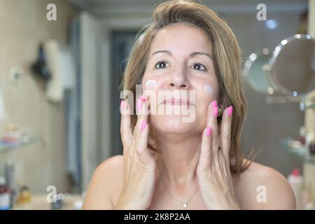 Reife weiße Frau mit Creme auf ihrem Gesicht zur Hautpflege und vor dem Spiegel im Badezimmer. Stockfoto