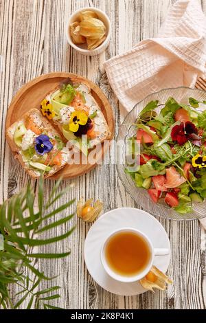 Pescetarian Food - Toast und Salat mit geräuchertem Lachs, Quark, essbaren Blumen, Gemüse und Gemüse. Tasse Tee. Ästhetische Herbstmittags Stockfoto