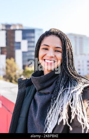 Fröhliche hispanische Hündin mit Dreadlocks, die mit einem Lächeln auf die Kamera blicken, während sie in der Nähe der roten Grenze des Gebäudes in der Stadt steht Stockfoto