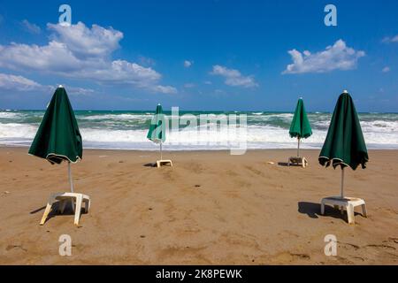 Grüne Sonnenschirme und weiße Plastiktische an einem einsamen Sandstrand mit Wellen, die im Hintergrund auf den Sand krachen, und blauem Himmel mit Wolken darüber. Stockfoto