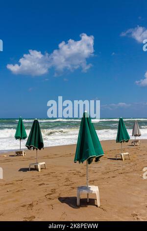 Grüne Sonnenschirme und weiße Plastiktische an einem einsamen Sandstrand mit Wellen, die im Hintergrund auf den Sand krachen, und blauem Himmel mit Wolken darüber. Stockfoto