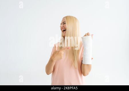 Porträt einer positiven blonden jungen Frau, die Zeigefinger auf einen gebrochenen Arm zeigt, eingewickelt in einen Gipsverband, der auf einem weißen isolierten Hintergrund steht. Stockfoto