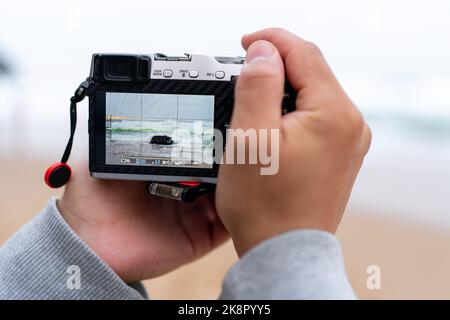 Reisefotoaufnahme mit spiegelloser Kamera in der Hand, Reiseblogger, Nahaufnahme von Männerhände mit spiegelloser Kamera, die beim Meerblick fotografiert Stockfoto