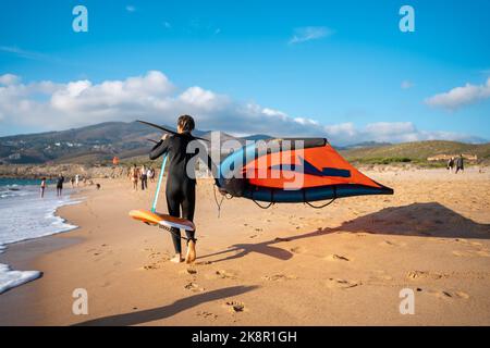 Foil Surfer beim Hydrofoil-Surfen im Meer an einem sonnigen Tag. Mann mit Kite-Tragflächenboot Ausrüstung zu Fuß Sandstrand Stockfoto