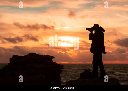 Portland, Dorset, Großbritannien. 24. Oktober 2022. Ein Fotograf trotzt dem wehenden Meereswind, um den Sonnenuntergang in Portland Bill, Dorset, an einem für die Saison unangenehmen Tag im Süden Englands einzufangen. Kredit: Peter Lopeman/Alamy Live Nachrichten Stockfoto