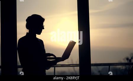 Dunkle Silhouette einer Geschäftsfrau, die etwas in den Laptop schreibt, es in den Händen hält, von einer Seite zur anderen um das Büro geht, vor dem Hintergrund eines großen Bürofensters, bei Sonnenuntergang, in den Lichtstrahlen. Hochwertige Fotos Stockfoto