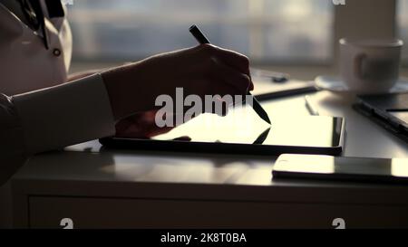 Dunkle Silhouette der Geschäftsfrau, Nahaufnahme der Hände. Sie schreibt etwas in Tablet, als nächstes ist ein Laptop auf dem Tisch. Unscharfer Fensterhintergrund , ein Lichtstrahl wird auf dem Tablet-Bildschirm reflektiert. Hochwertige Fotos Stockfoto