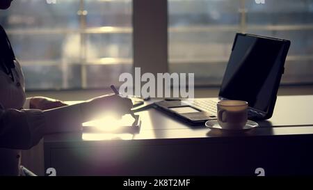 Dunkle Silhouette der Geschäftsfrau, Nahaufnahme der Hände. Sie schreibt etwas in Tablet, als nächstes ist ein Laptop auf dem Tisch. Unscharfer Fensterhintergrund , ein Lichtstrahl wird auf dem Tablet-Bildschirm reflektiert. Hochwertige Fotos Stockfoto