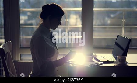 Dunkle Silhouette der jungen fokussierten aufmerksamen Geschäftsfrau trägt Brille, schreibt etwas in Tablet, als nächstes ist ein Laptop auf dem Tisch. Gegen das Fenster, ein Lichtstrahl wird auf dem Tablet-Bildschirm reflektiert. Hochwertige Fotos Stockfoto