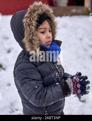 Porträt des kleinen Latino-Jungen im Profil mit besorgtem Gesicht und umklammernden Händen, im Winter im Schnee, in einem pelzigen Kapuzenmantel Stockfoto