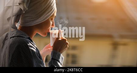 Banner der jungen schönen Frau trinkt heißen dampfenden Kaffee oder Tee im Sonnenschein Licht auf dem Balkon im Hotel oder zu Hause. Frisches Mädchen nach der Dusche tragen Stockfoto
