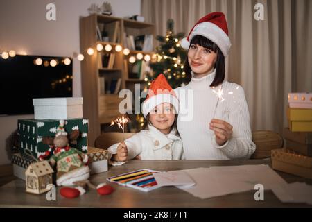 Fröhliches nettes Mädchen und ihre Mutter mit charmanten Lächeln in Santa Hüte sitzen auf dem Tisch mit funkelt auf dem Hintergrund von Weihnachtsbaum und dekorierten Wohnzimmer. Stockfoto