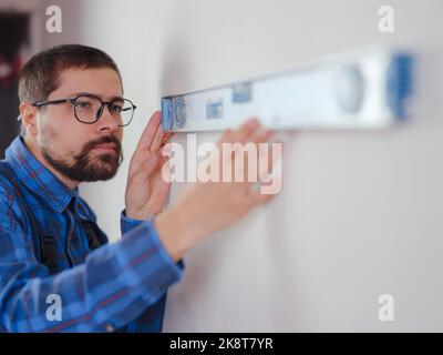 Junger Mann in blau Arbeitsanzug tun Reparatur Wohnung. Renovierungskonzept für das Haus. Kurzer Schuss eines hübschen bärtigen jungen Mannes, der zu Hause ein Level-Werkzeug hält Stockfoto