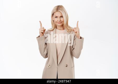 Blonde Frau mittleren Alters mit skeptischem Gesicht, mit den Fingern nach oben zeigend, Zweifel am Produkt habend, über weißem Hintergrund stehend Stockfoto