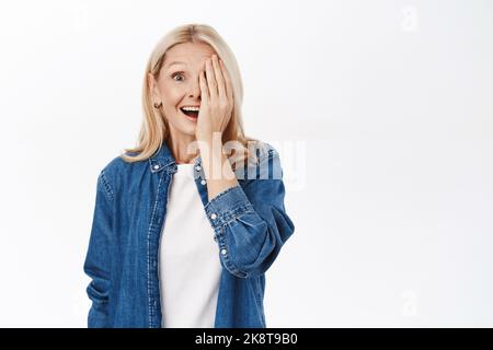 Porträt einer schönen älteren Frau mit blonden Haaren, bedeckt die Hälfte des Gesichts, eine Seite, lächelnd und schauend auf die Kamera glücklich, stehend über weißem Hintergrund Stockfoto