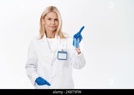 Skeptische Ärztin, unzufrieden, zeigt oben rechts mit unzufriedenem, zweifelhaftem Gesichtsausdruck, steht auf weißem Hintergrund Stockfoto