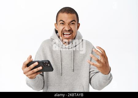 Wütend Mann reagieren auf smeth auf Smartphone, schreiend und zitternd wütend, halten Handy in der Hand, stehen über weißem Hintergrund Stockfoto