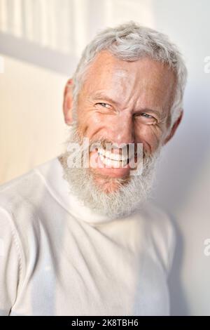Lächelnder bärtiger älterer Mann, der lachend an der weißen Wand im Sonnenlicht steht. Stockfoto