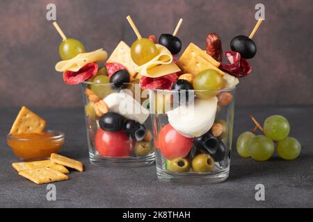 Jarcuterie in zwei Glas umfasst verschiedene Arten von Wurst, Gurken, Käse, Nüsse, Gemüse und Obst. Moderne Version von festlichem Snack auf dunklem Rücken Stockfoto