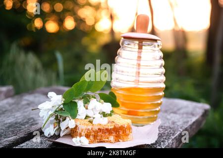 Glas Akazienhonig im Hintergrund des Sonnenuntergangs. Ein Wabenblatt liegt auf Handwerkspapier auf Holzbrettern. Robinienhonig mit Blütenakazie blüht im Sommer Stockfoto