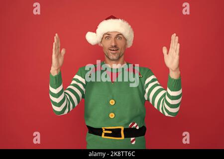 Staunender Mann im Elf Kostüm. weihnachtsmann im weihnachtsmann Hut auf rotem Hintergrund. Frohes neues Jahr. Stockfoto