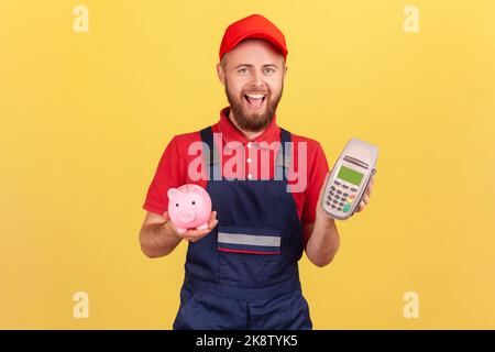 Fröhlicher bärtiger Arbeiter Mann hält in der Hand POS kontaktloses Zahlungsterminal und Sparschwein, Cashback, mit paypass für Service-Zahlung. Innenaufnahme des Studios isoliert auf gelbem Hintergrund. Stockfoto
