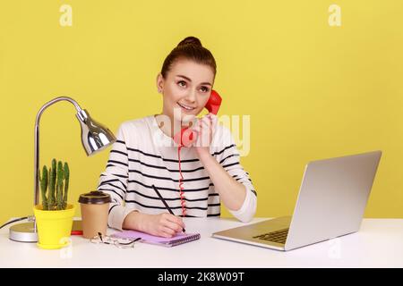 Porträt einer lächelnden Frau, die auf einem roten Retro-Telefon spricht, mit verträumtem Gesichtsausdruck wegschaut, am Arbeitsplatz sitzt und in einem Notizblock schreibt. Studio-Innenaufnahme isoliert auf gelbem Hintergrund. Stockfoto