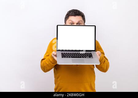 Porträt eines Mannes, der die Hälfte des Gesichts mit einem Laptop mit leerem Display für Werbung oder Werbetexte bedeckt und einen Hoodie im urbanen Stil trägt. Innenaufnahme des Studios isoliert auf weißem Hintergrund. Stockfoto