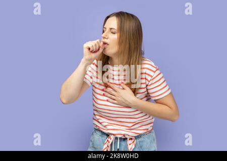 Porträt einer kranken, ungesunden, blonden Frau mit gestreiftem T-Shirt, die Husten und Schmerzen in der Lunge, Erkältung und Grippesymptome hat. Innenaufnahme des Studios isoliert auf violettem Hintergrund. Stockfoto