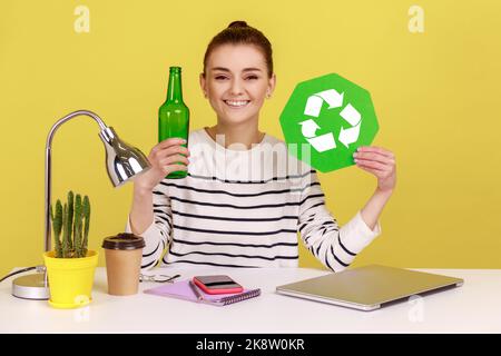 Verantwortungsbewusste selbstbewusste glückliche Frau mit grünem Recycling-Schild in der Hand und Glasflasche, sitzend am Arbeitsplatz mit Laptop. Studio-Innenaufnahme isoliert auf gelbem Hintergrund. Stockfoto