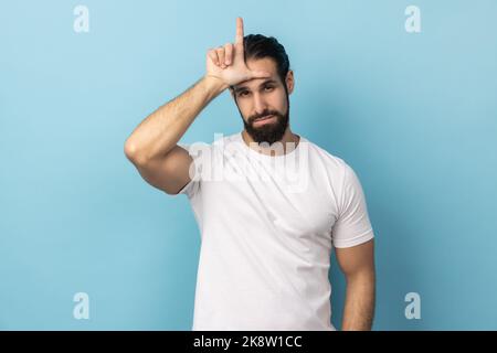 Ich habe meinen Job verloren. Porträt eines Mannes mit Bart und weißem T-Shirt mit einer loseren Geste, L-Finger-Schild auf der Stirn, verärgert über Entlassung, unglücklicher Tag. Innenaufnahme des Studios isoliert auf blauem Hintergrund. Stockfoto