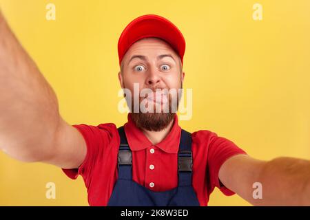 Kindlicher Arbeiter mit roter Mütze und blauer Uniform, der Selfie macht, die Kamera anschaut und die Zunge aus dem POV zeigt, aus der Perspektive des Fotos. Innenaufnahme des Studios isoliert auf gelbem Hintergrund. Stockfoto