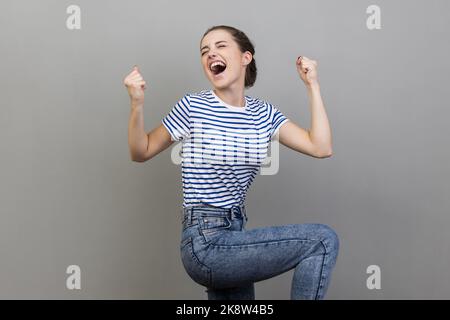 Ich bin Champion. Porträt einer Frau im gestreiften T-Shirt, die vor Freude schreit, ihre Fäuste hochhebt und den erfolgreichen Sieg feiert. Innenaufnahme des Studios isoliert auf grauem Hintergrund. Stockfoto