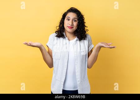 Ich bin mir nicht sicher. Porträt einer zweideutigen Frau mit dunklen welligen Haaren, die mit erhobenen Armen steht, wegschaut und nicht weiß, was zu tun ist. Innenaufnahme des Studios isoliert auf gelbem Hintergrund. Stockfoto