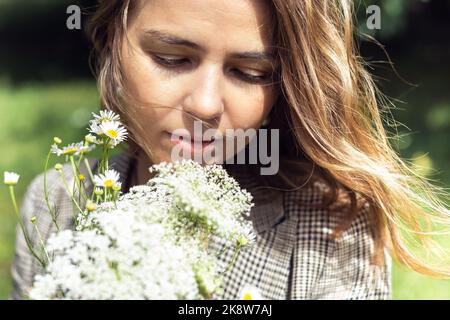 Nahaufnahme blonde, ruhige blonde Frau, die einen Strauß von Feldblumen hält und ihn riecht. Aromatherapie oder Allergie. Naturpark Stockfoto