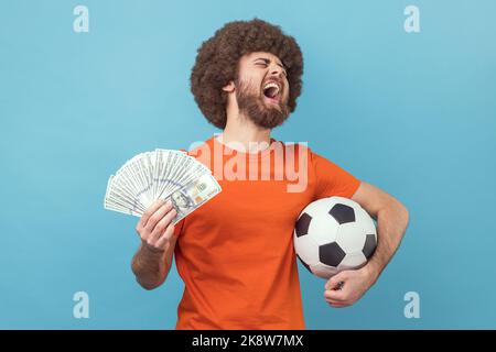 Aufgeregter Mann mit Afro-Frisur in orangefarbenem T-Shirt mit Fußballball und hundert Dollar-Scheinen, schreiend vor Glück, Wetten und Gewinnen. Innenaufnahme des Studios isoliert auf blauem Hintergrund. Stockfoto